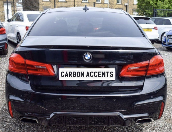 A black BMW with visible carbon accents is parked on gravel among other cars. The license plate reads "CARBON ACCENTS," emphasizing the car's design features.
