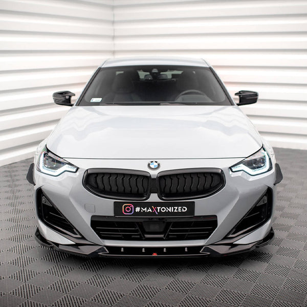 A white BMW car is stationary in a minimalist room with geometric walls and flooring. The front grille and headlights are prominent. Text on the car reads "#MAXTONIZED."