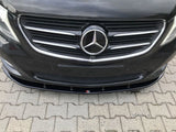 A black car front features a prominent emblem in the center of a chrome grille, parked on a paved surface with interlocking bricks. The license plate area is empty.