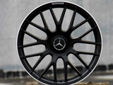 A black alloy wheel with a Mercedes emblem rests on a concrete block outdoors. The rim features 