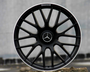 A black alloy wheel with a Mercedes emblem rests on a concrete block outdoors. The rim features "AMG" text at the top edge, and the background is a blurred gray wall.