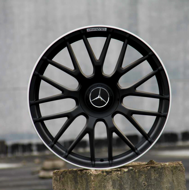 A black alloy wheel with a Mercedes emblem rests on a concrete block outdoors. The rim features "AMG" text at the top edge, and the background is a blurred gray wall.