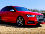 A red Audi car is parked on a gravel path surrounded by greenery and a wooden fence under a clear blue sky.