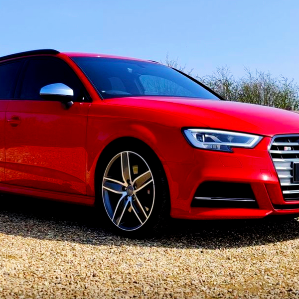 A red Audi car is parked on a gravel path surrounded by greenery and a wooden fence under a clear blue sky.