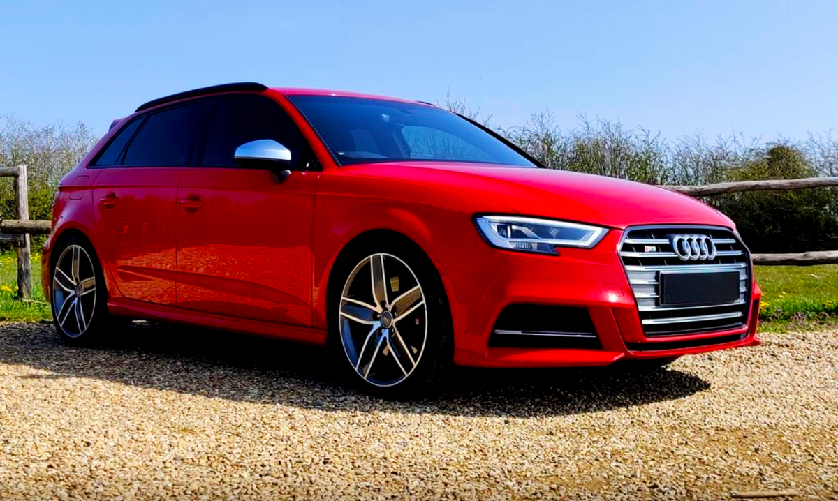 A red Audi car is parked on a gravel path surrounded by greenery and a wooden fence under a clear blue sky.