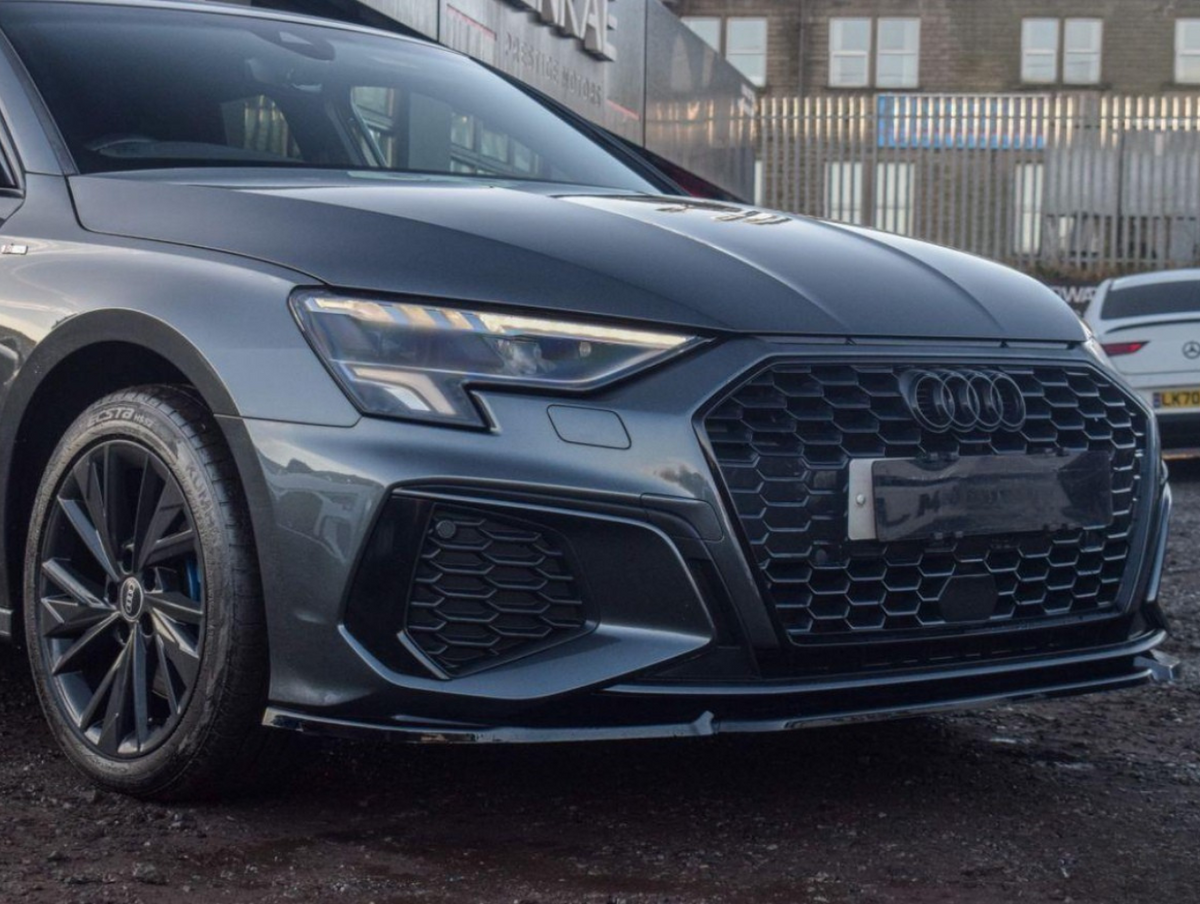 A sleek, dark Audi car is parked on a gravel lot. Its hexagonal grille and LED headlights are prominent. In the background, there’s a building and other parked vehicles.