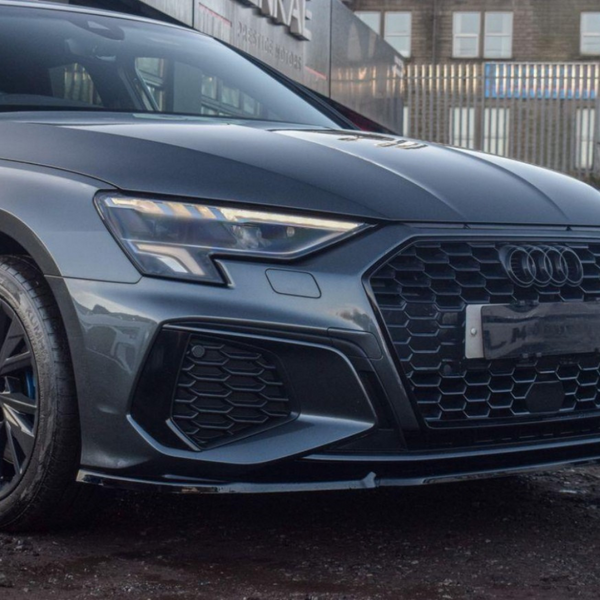 A sleek, dark Audi car is parked on a gravel lot. Its hexagonal grille and LED headlights are prominent. In the background, there’s a building and other parked vehicles.