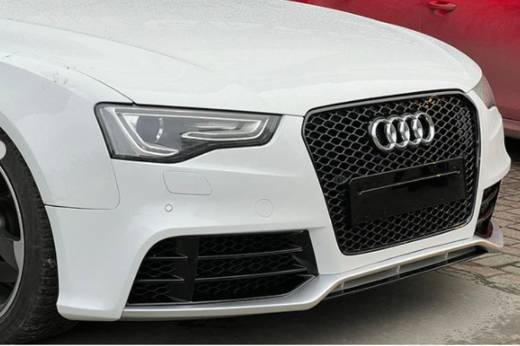 A white car's front features a prominent grille with the Audi logo. It's parked on a paved surface, next to a red car visible in the background.