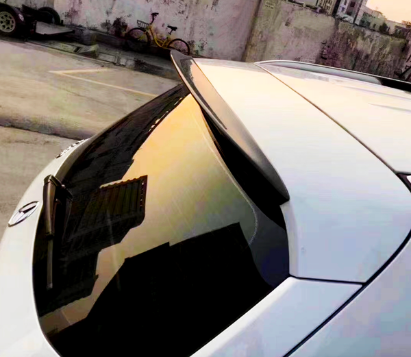 Car rear window reflects urban sunset; rooftop spoiler above, parking lot and bicycle against a concrete wall in the background.
