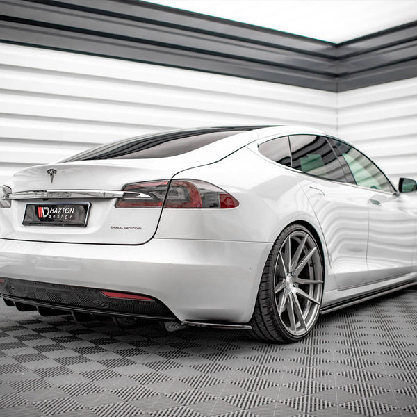 A white Tesla sedan parked in a modern showroom with checkered flooring and paneled walls. License plate reads "MAXTON design." The car has large, silver alloy wheels and tinted windows.