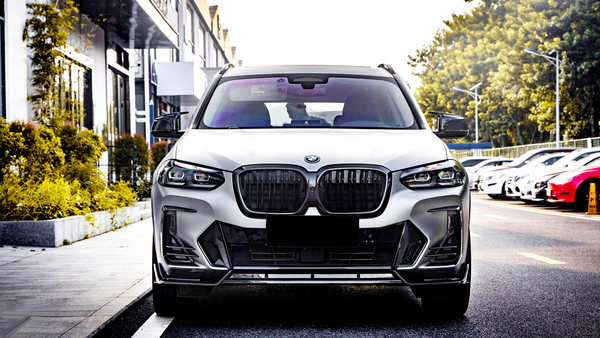 A white car is parked on a street, facing forward. It is surrounded by parked cars and trees line the side of a nearby building.