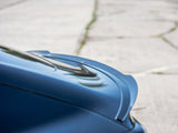 A sleek, blue car spoiler is angled upward against a blurred background of a paved surface, suggesting a focus on aerodynamics and style in a parking area.