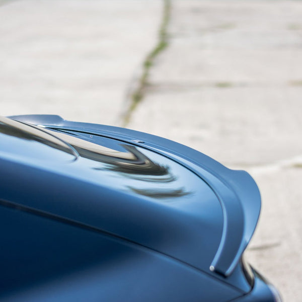A sleek, blue car spoiler is angled upward against a blurred background of a paved surface, suggesting a focus on aerodynamics and style in a parking area.