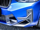A blue car's front bumper with carbon fiber accents, features a grille and headlight, parked on a paved surface. The tire shows some white specks.