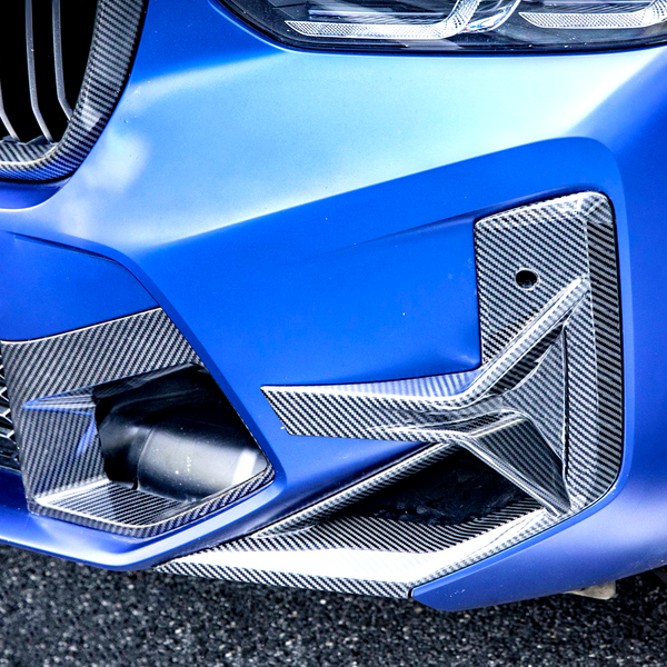 A blue car's front bumper with carbon fiber accents, features a grille and headlight, parked on a paved surface. The tire shows some white specks.