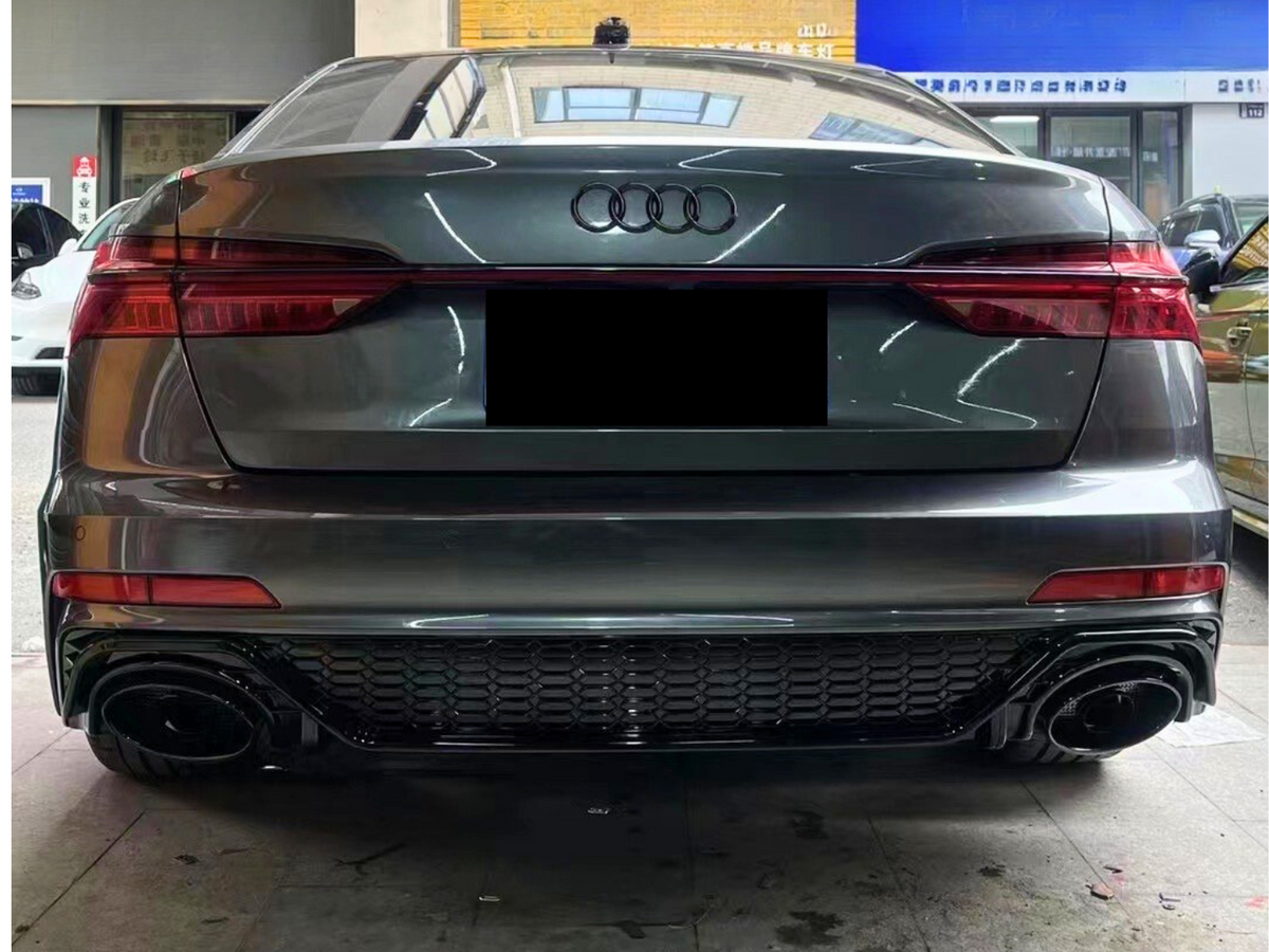 A gray Audi car is parked indoors, seen from the rear, showcasing its taillights and exhaust pipes. In the background, other cars and a blue signboard with Chinese text are visible.
