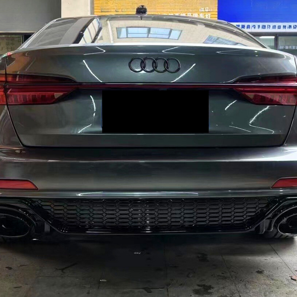 A gray Audi car is parked indoors, seen from the rear, showcasing its taillights and exhaust pipes. In the background, other cars and a blue signboard with Chinese text are visible.
