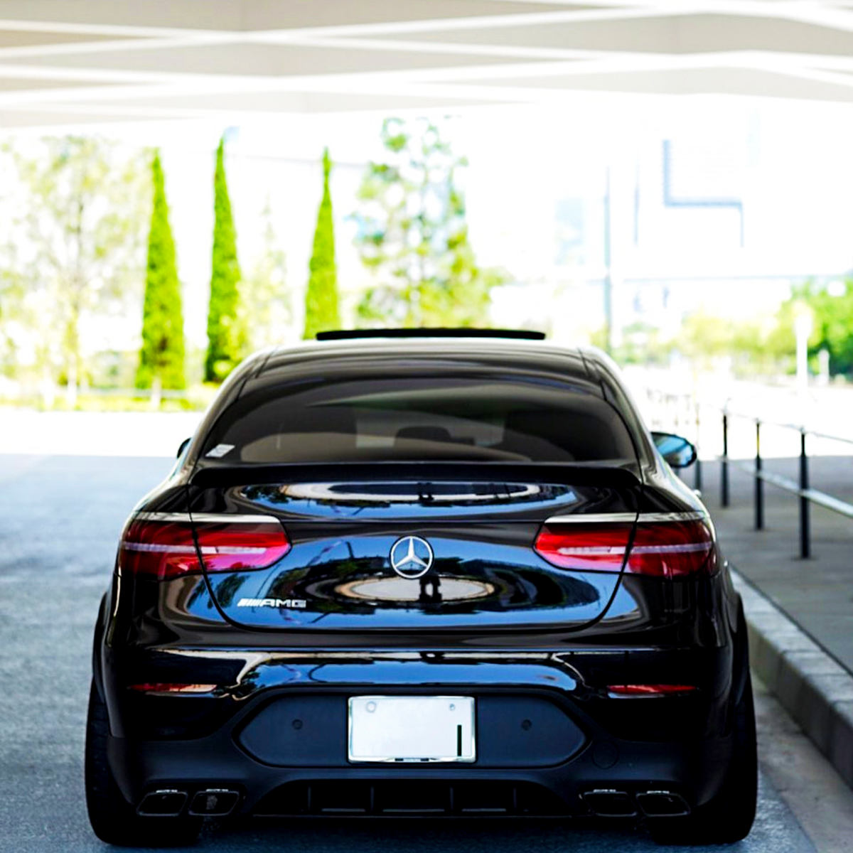 A black Mercedes-Benz parked under a modern, white structure with geometric patterns, surrounded by trees and urban scenery. The rear license plate is visible but unreadable.