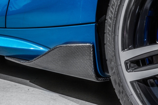 A blue car with a carbon fiber side skirt is parked, featuring a detailed close-up of its wheel and tire on a smooth concrete surface.