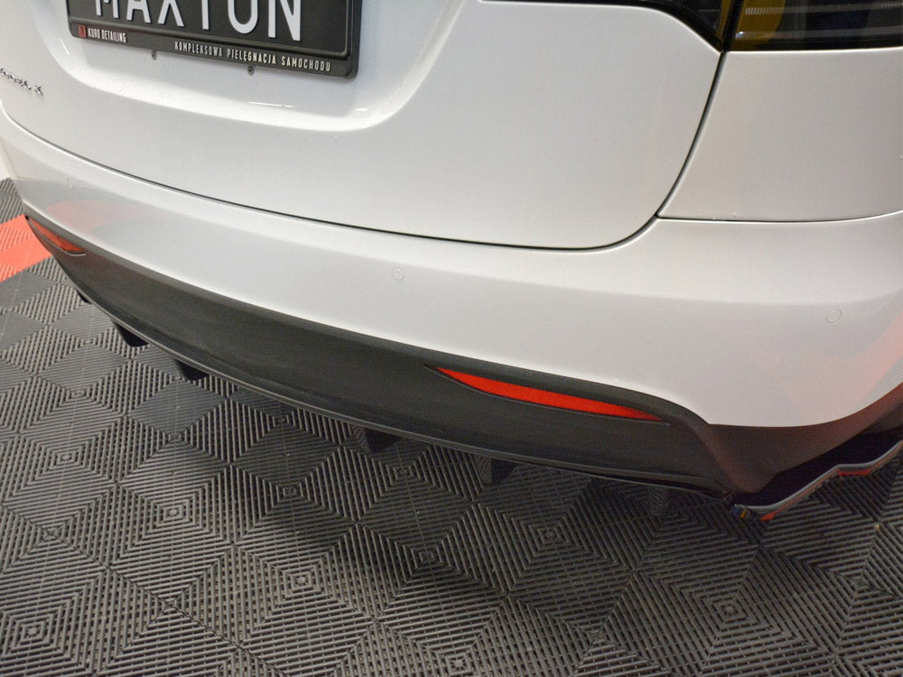 White car's rear with a black bumper and diffuser, displaying a license plate reading "MAXTON." The car is parked on a patterned floor in a showroom.