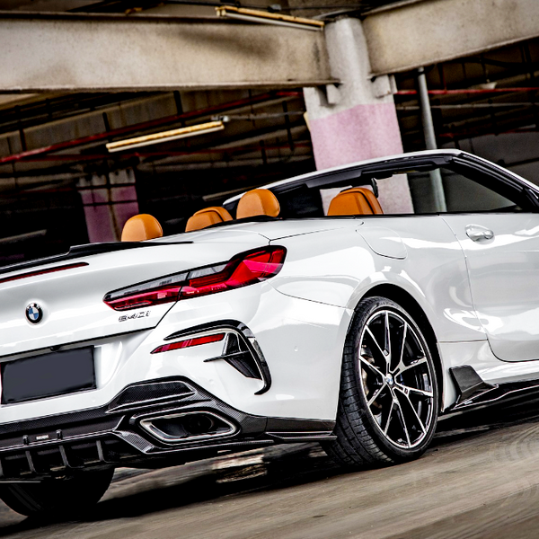 A white convertible car, with top down and orange seats visible, is parked in an empty, dimly-lit parking garage. The car has twin exhausts and modern alloy wheels.