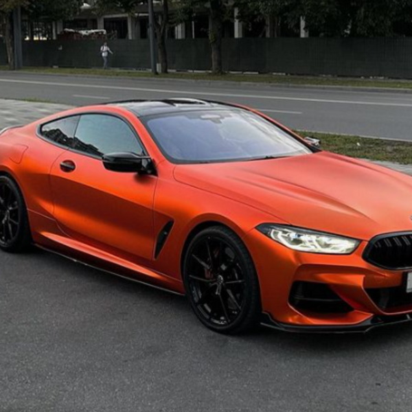 A sleek, orange sports car is parked beside a road, set against a backdrop of trees and an urban environment.
