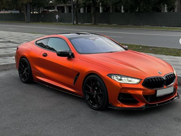 A sleek, orange sports car is parked beside a road, set against a backdrop of trees and an urban environment.