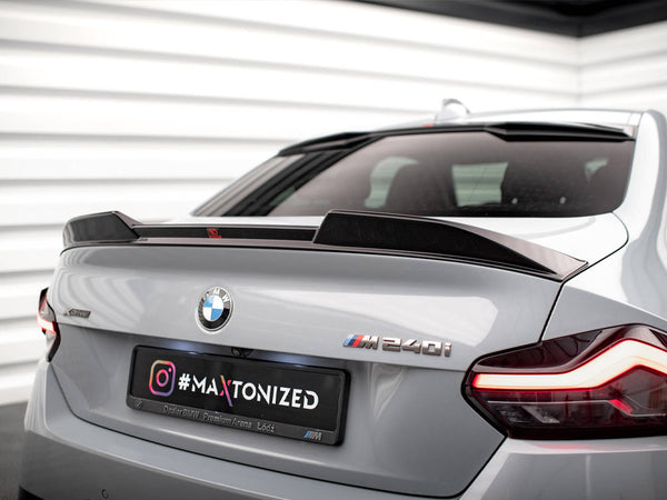 A silver BMW M240i displays a custom spoiler on a parking deck. The badge and taillights are visible. A license plate reads: "#MAXTONIZED Instagram, Dealer BMW Premium Arena Łódź." Walls feature white panels.