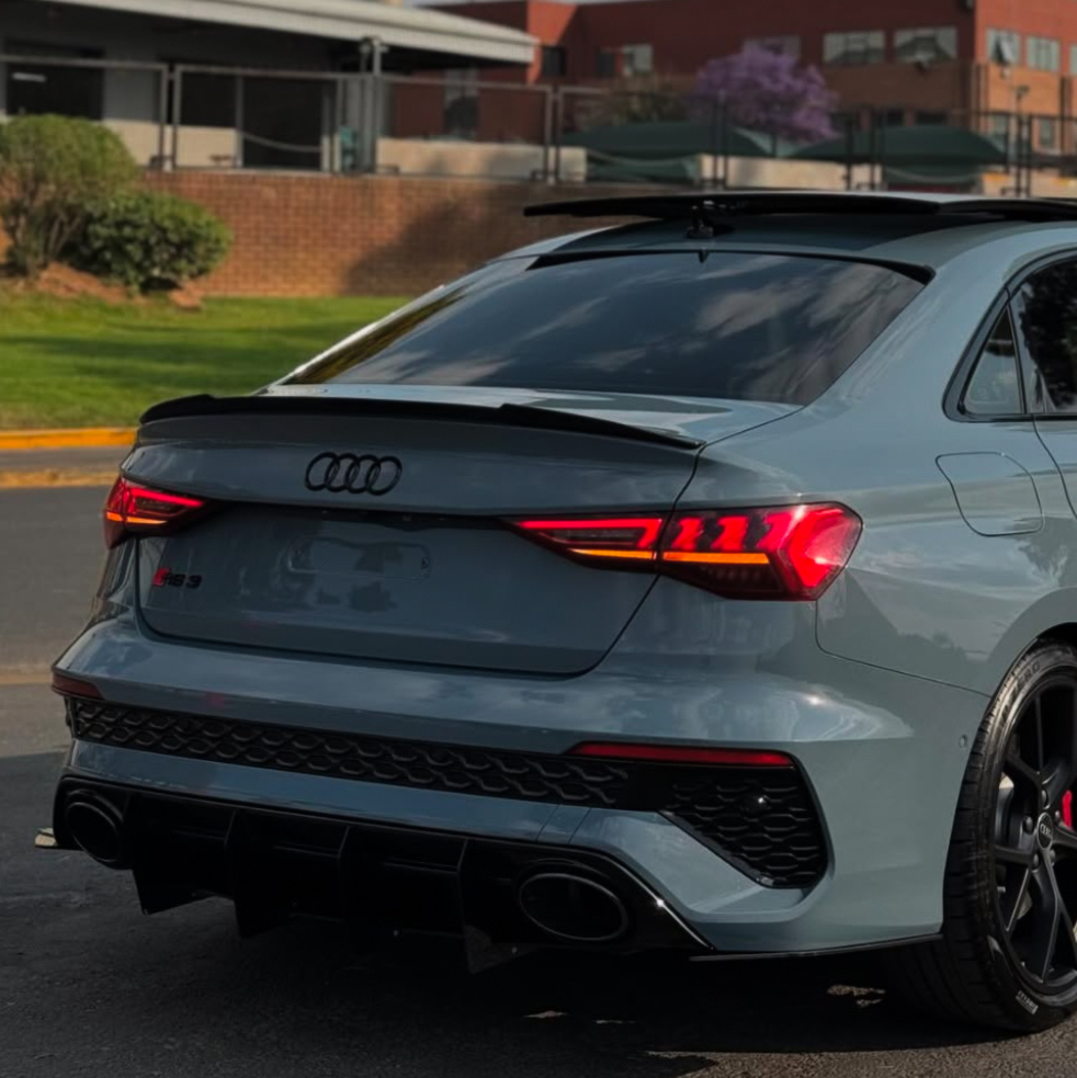 A gray Audi RS3 parked, featuring illuminated taillights and dual exhausts, in a paved area with green shrubs and brick buildings in the background.