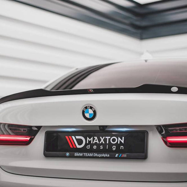 A white BMW car, featuring red tail lights, is parked indoors. The license plate reads "MAXTON design, BMW TEAM Długołęka" in a sleek, modern garage.