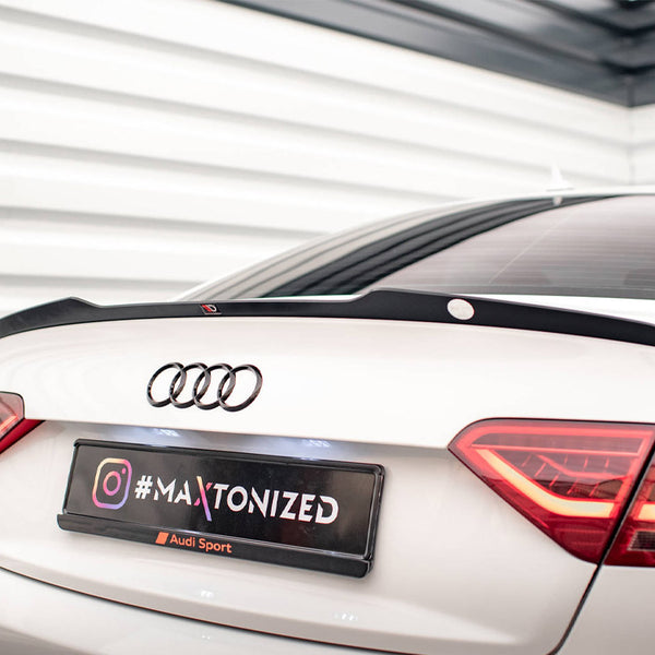 A white Audi car with a spoiler is parked in a corner with white geometric walls. The license plate reads "#MAXTONIZED" with an Instagram logo and "Audi Sport" below.