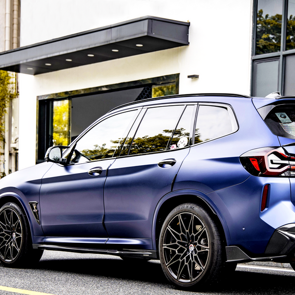 A sleek, metallic blue SUV is parked on a street, featuring intricate black alloy wheels, against a backdrop of a modern white building with large windows and greenery reflected in them.