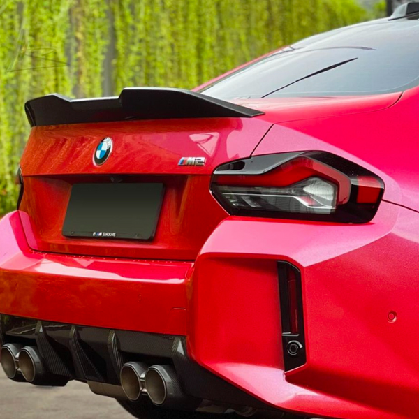 A red sports car with a visible quad exhaust system and a rear spoiler is parked on a paved surface, adjacent to a wall covered with vertical greenery. Text: "BMW" logo, "M2".