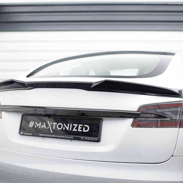 A white Tesla car is parked, showcasing a black spoiler on its rear. The license plate reads "#MAXTONIZED SERWIS POJAZDÓW ELEKTRYCZNYCH TEL +48 660 323 329" in a garage setting.