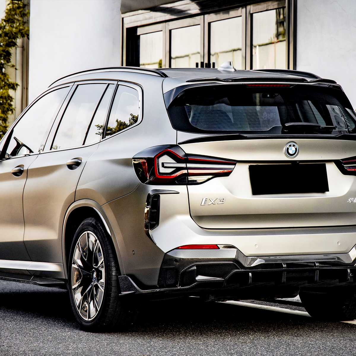 A silver SUV is parked on a city street, with surrounding greenery and modern glass buildings. The license plate and model name "iX3" are visible on the rear.