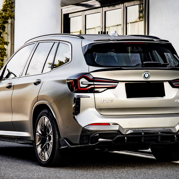 A silver SUV is parked on a city street, with surrounding greenery and modern glass buildings. The license plate and model name "iX3" are visible on the rear.
