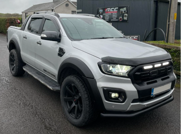 A white pickup truck with large tires and prominent front grille sits parked on a wet road. Behind, there's a dark building with a banner that reads "ECC" and lists automotive services.