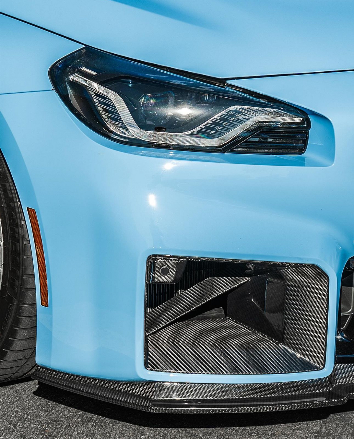 A car's blue front end features an angular headlight and a carbon fiber air intake, with a smooth surface and tire visible, suggesting a sleek, sporty design parked on asphalt.