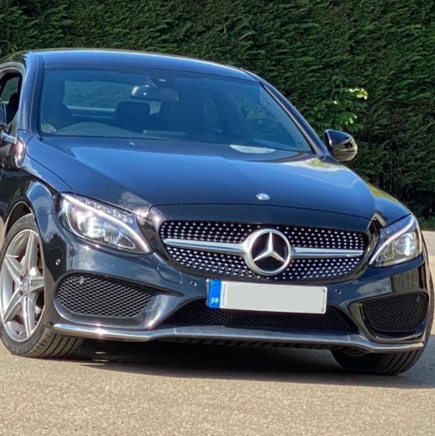 A sleek, black Mercedes-Benz car is parked on a sunlit road, surrounded by lush greenery. The car's distinctive grille and emblem are prominently displayed. No visible text on the car.