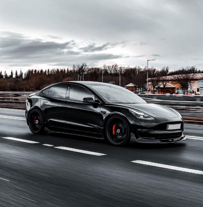 A sleek black car moves swiftly along a highway, surrounded by blurred motion, trees, and buildings under a cloudy sky, emphasizing speed and modern design.