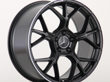 A black alloy car wheel features a multi-spoke design with the Mercedes-Benz emblem in the center. The AMG logo is written on the rim. It sits on a light surface.