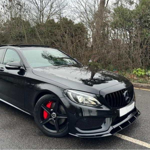 A black car with black grille and red brake calipers is parked on a wet road, surrounded by bare trees and shrubbery.