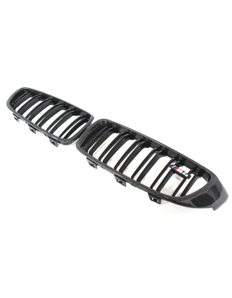Black car grille with vertical slats and a small "M2" badge rests on a plain white background, emphasizing its glossy carbon fiber texture and modern design.