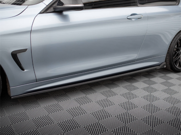 A metallic silver car with sleek side skirts is parked on a geometric-patterned floor, emphasizing its modern design and low profile. The car's door and wheel are partially visible.