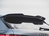 A sleek car rear spoiler extends over the tinted back window of a gray vehicle, with the Audi logo visible, set against a cloudy sky.
