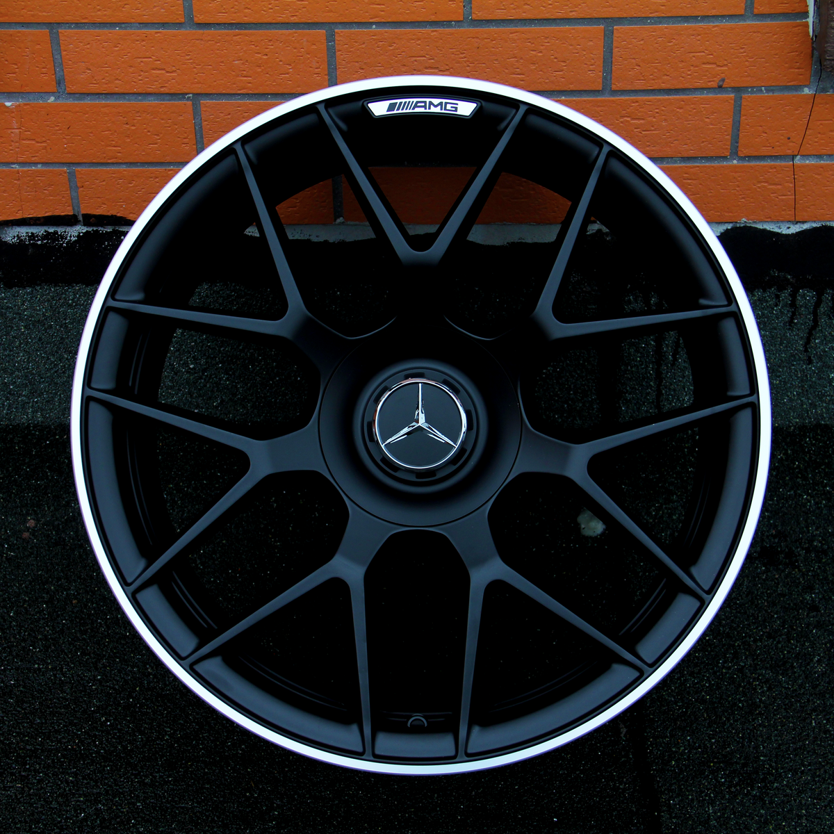 A black alloy wheel with a Mercedes-Benz logo and "AMG" branding is placed upright against an orange brick wall on a textured black surface.