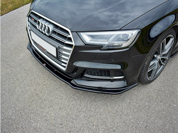 A black car, viewed from the front, is parked on a paved surface. It features a prominent grille with a logo and sleek headlights, with grass visible in the background.