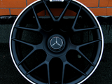 A black alloy wheel with a central Mercedes-Benz logo and AMG branding rests against an orange brick wall on a dark surface, showcasing an intricate spoke design.