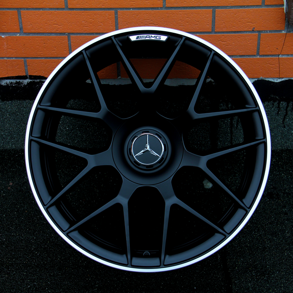 A black alloy wheel with a central Mercedes-Benz logo and AMG branding rests against an orange brick wall on a dark surface, showcasing an intricate spoke design.