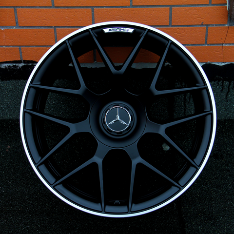 A black alloy wheel with a central Mercedes-Benz logo and AMG branding rests against an orange brick wall on a dark surface, showcasing an intricate spoke design.